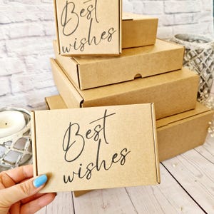 May include: A collection of small, brown cardboard boxes, some stacked. The top box and the box being held display the words "Best wishes" in black script. The boxes are on a light surface with a brick wall background.