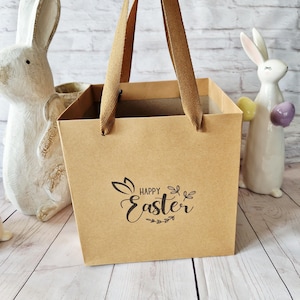 May include: A brown paper gift bag with brown handles, printed with "Happy Easter" in black. Two ceramic Easter bunny ornaments are visible in the background.