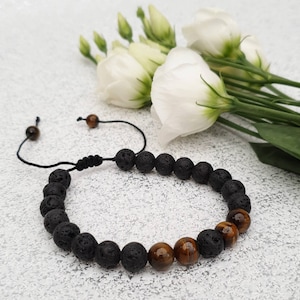 May include: A black lava stone bracelet with brown tiger eye beads. The bracelet is adjustable with a black cord and features a sliding knot closure. White flowers are in the background.