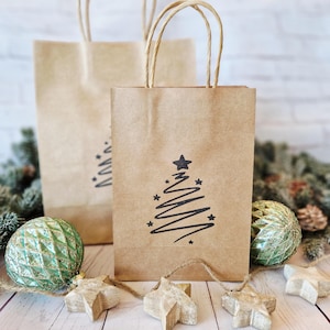 May include: Two brown paper gift bags with twisted paper handles. One bag features a black Christmas tree design with stars. The bags are on a wooden surface with Christmas ornaments and star-shaped decorations.
