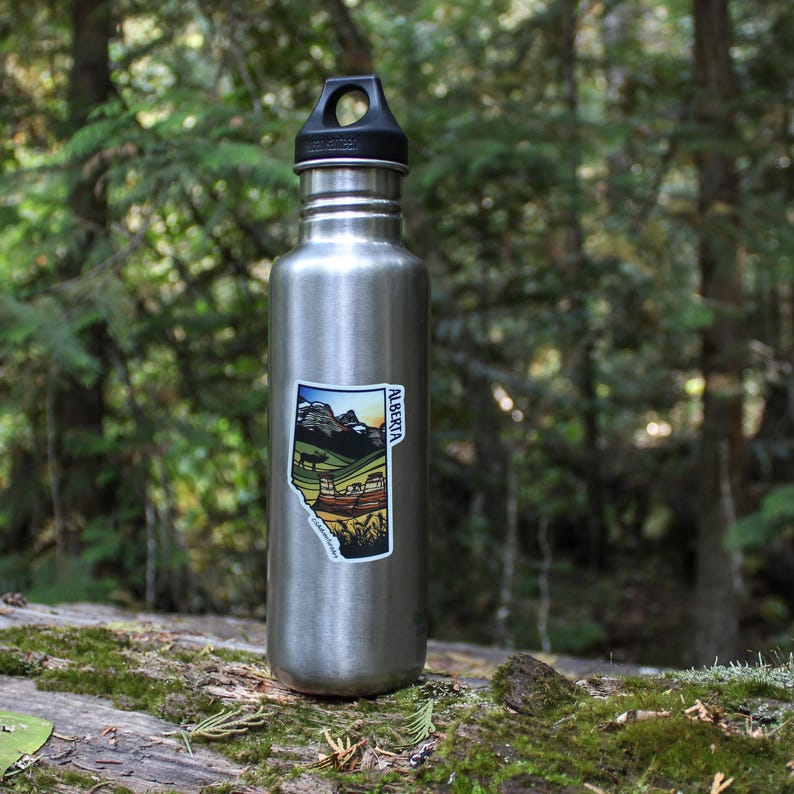 Beautiful waterproof sticker depicting Banff National Park applied to a water bottle in a mossy forest