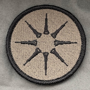 May include: Embroidered circular patch with a tan centre and a black sunburst design. The sunburst has eight pointed elements. The patch is edged with black stitching. The background is a muted green fabric.