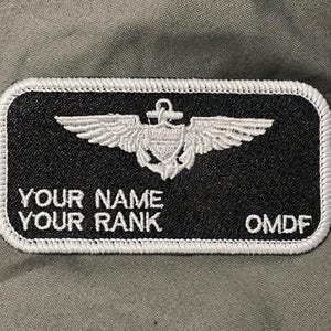 Peut inclure: Un écusson rectangulaire noir avec du texte brodé blanc et une bordure blanche. Le texte indique "YOUR NAME", "YOUR RANK" et "OMDF". Un emblème blanc d'ailes et d'une ancre est centré au-dessus du texte.