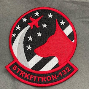 Puede incluir: Parche bordado con una silueta de perro rojo, un avión y el texto "STRKFITRON-132". El parche tiene un fondo negro con estrellas blancas y un borde rojo. El avión es rojo y el perro es de color rojo liso.