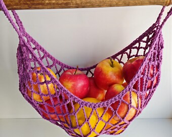 May include: A handmade, purple crochet fruit hammock filled with red and yellow apples. The hammock is suspended from a wooden rod, creating a decorative and functional storage solution for the kitchen.