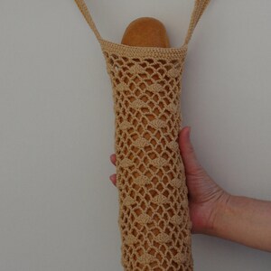 May include: A beige, crocheted bread bag, designed to hold a loaf of bread. The bag is made of a net-like pattern, with a rounded bottom and a strap for carrying. The bag is hanging from a metal hook.
