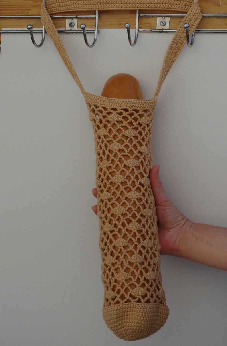 May include: A beige, crocheted bread bag, designed to hold a loaf of bread. The bag is made of a net-like pattern, with a rounded bottom and a strap for carrying. The bag is hanging from a metal hook.