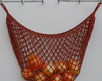 May include: A rust-colored, hand-crocheted net bag with a diamond-shaped mesh pattern, suspended from a metal rack. The bag contains several yellow onions, visible through the open weave of the yarn, which forms a wide, flexible storage pouch.