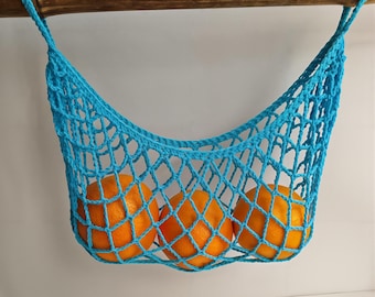 May include: A turquoise crocheted net bag, suspended from a wooden rod, holds three bright orange oranges. The bag's open weave design allows visibility of the fruit. The bag is a vibrant accent.
