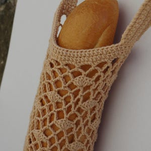 May include: A beige, crocheted bread bag holds a golden-brown baguette. The bag has a net-like design with two handles. The bread bag is designed to carry a loaf of bread.