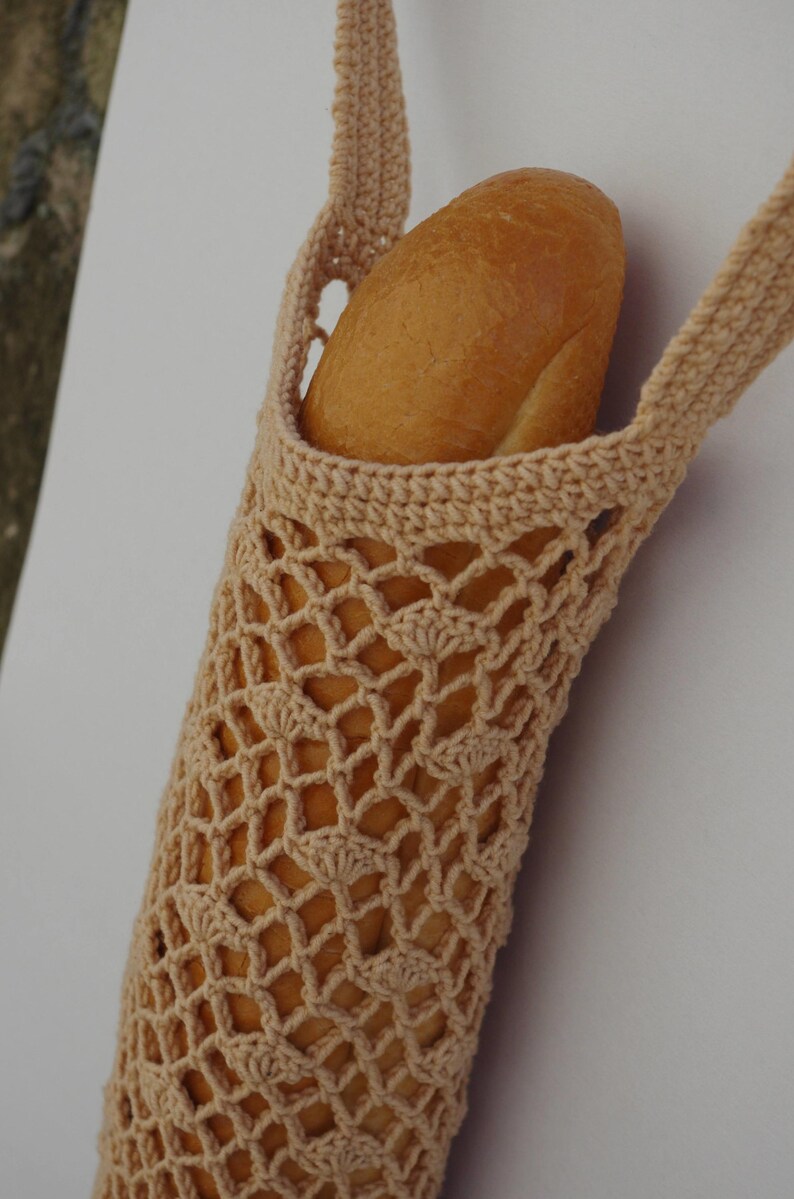 May include: A beige, crocheted bread bag holds a golden-brown baguette. The bag has a net-like design with two handles. The bread bag is designed to carry a loaf of bread.