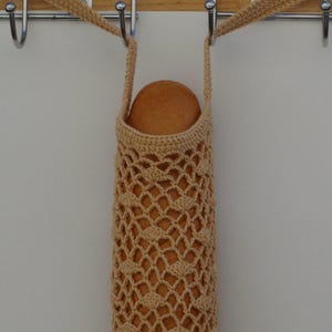 May include: A beige, crocheted bread bag with a lattice design, holding a round loaf of bread. The bag has a strap for carrying and is hanging from a metal hook. The bag is made of a soft, natural fiber.