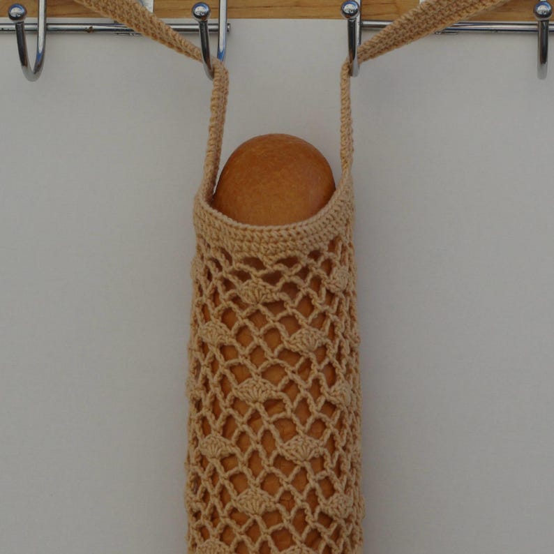 May include: A beige, crocheted bread bag with a lattice design, holding a round loaf of bread. The bag has a strap for carrying and is hanging from a metal hook. The bag is made of a soft, natural fiber.