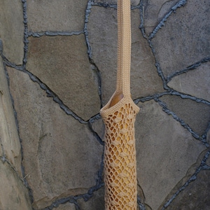 May include: A beige, crocheted bag with a long handle, designed to hold a baguette. The bag has an open, net-like weave, allowing a glimpse of the bread inside. The handle is held up by a hand against a stone wall.
