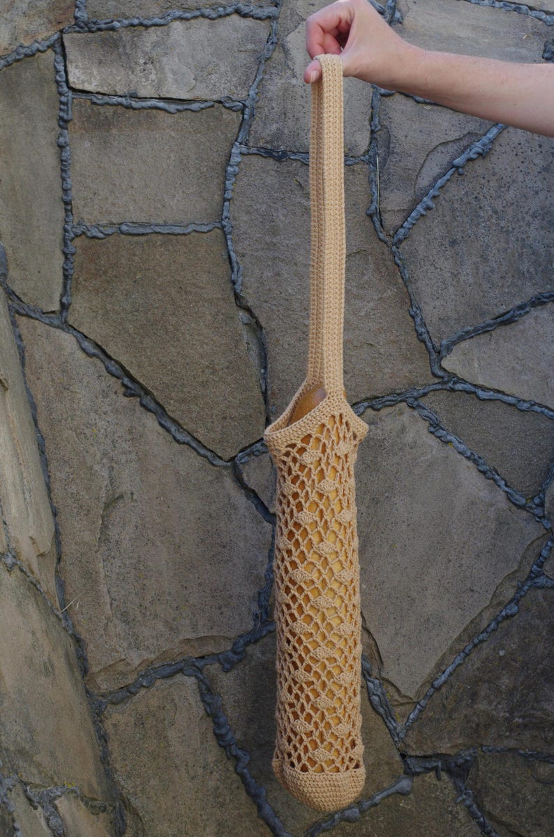May include: A beige, crocheted bag with a long handle, designed to hold a baguette. The bag has an open, net-like weave, allowing a glimpse of the bread inside. The handle is held up by a hand against a stone wall.