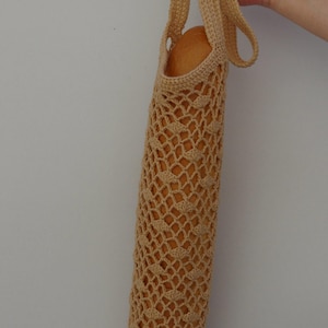 May include: A beige crocheted baguette bag, held by a hand. The bag is cylindrical with a diamond-patterned mesh design. The bag is filled with a loaf of bread. The bag has a handle for carrying.