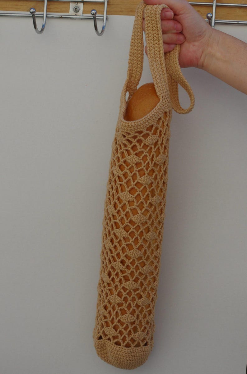 May include: A beige crocheted baguette bag, held by a hand. The bag is cylindrical with a diamond-patterned mesh design. The bag is filled with a loaf of bread. The bag has a handle for carrying.