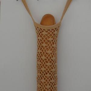 May include: A beige, crocheted bread bag, hanging from a metal hook. The bag is filled with a loaf of bread. The bag has a long, cylindrical shape with a diamond-patterned weave. The bag is suspended by two straps.
