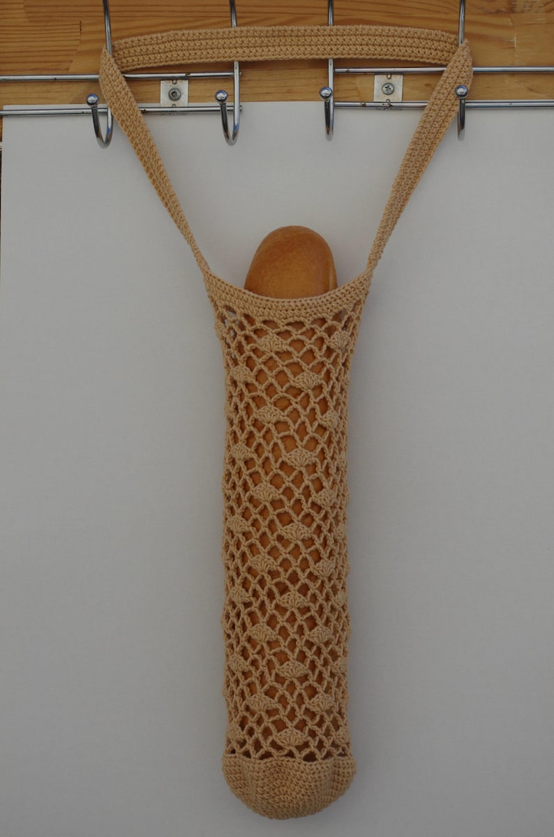 May include: A beige, crocheted bread bag, hanging from a metal hook. The bag is filled with a loaf of bread. The bag has a long, cylindrical shape with a diamond-patterned weave. The bag is suspended by two straps.