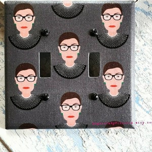 May include: A gray light switch cover with a repeating pattern of a woman with black hair, glasses, and a black lace collar. The woman's face is pink and she is wearing green earrings. The text "DesignsByPrintables etsy.com" is in the bottom right corner.