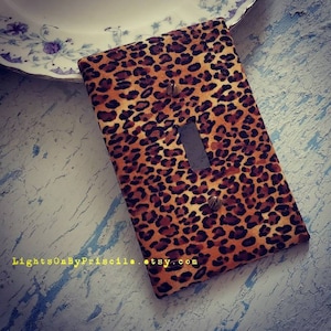 May include: A light switch cover with a leopard print design. The cover is brown and orange with black spots. The switch plate is made of fabric and has a light switch cutout in the centre.