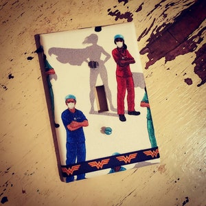 May include: A white light switch cover with a Wonder Woman logo at the bottom. The cover features a pattern of healthcare workers wearing masks and scrubs, with a superhero silhouette in the background.