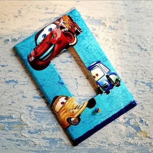 May include: A rectangular picture frame featuring characters from the animated film "Cars." The frame has a light blue background with a repeating pattern and a dark blue border. The characters Lightning McQueen, Mater, and others are printed on the fabric.