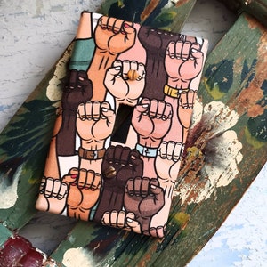 May include: A light switch cover featuring a colourful pattern of raised fists in various skin tones. The fists are all clenched and the pattern is repeated across the entire surface of the cover.
