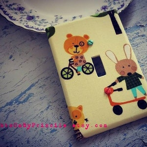 May include: A light yellow light switch cover with a brown bear riding a bicycle and a brown rabbit pushing a red wagon. The background is a green and yellow pattern with trees and leaves. The text "lightsOnByPriscilla etsy.com" is at the bottom of the cover.
