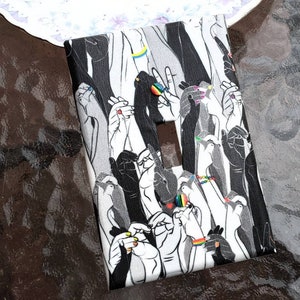 May include: A light switch cover with a black and white design of many hands reaching up. The hands are all different sizes and shapes, and some are holding rainbow-colored bracelets. The design is a celebration of diversity and inclusion.