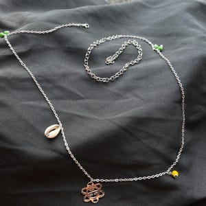 May include: Silver body chain necklace featuring a seashell pendant, a flower-shaped pendant, and small green and yellow beads. The necklace is shown against a dark backdrop.