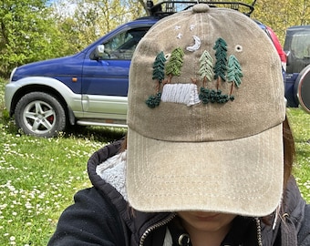 Könnte beinhalten: Eine beige Baseballkappe mit einem gestickten Design aus Mond, Sternen und Bäumen. Die Stickerei ist in Grün-, Weiß- und Brauntönen gehalten.