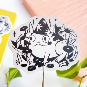 May include: Black and white sticker featuring a frog wearing a pointed hat, surrounded by flowers and sparkles. The frog is smiling and has large eyes.