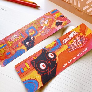 May include: Two colorful bookmarks featuring black cats and stacks of books. One bookmark has the text "you are on this side!" printed on it.