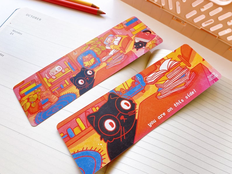 May include: Two colorful bookmarks featuring black cats and stacks of books. One bookmark has the text "you are on this side!" printed on it.