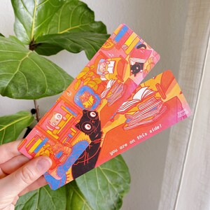 May include: Two colorful bookmarks with a cat and book illustration. The bookmark on the left has a black cat with a book in its paws. The bookmark on the right has a stack of books and the text "You are on this side!"