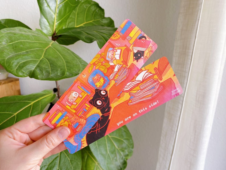 May include: Two colorful bookmarks with a cat and book illustration. The bookmark on the left has a black cat with a book in its paws. The bookmark on the right has a stack of books and the text "You are on this side!"