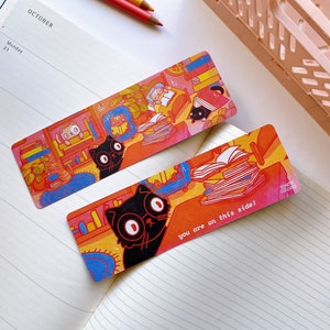 May include: Two colorful bookmarks featuring black cats reading books. The bookmark on the left has a cat with glasses reading a book in a bookshelf. The bookmark on the right has a cat with big eyes reading a stack of books. The text on the bookmark reads "you are on this side!"