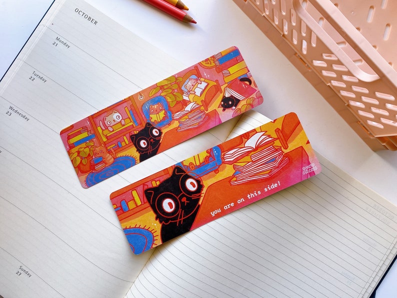 May include: Two colorful bookmarks featuring black cats reading books. The bookmark on the left has a cat with glasses reading a book in a bookshelf. The bookmark on the right has a cat with big eyes reading a stack of books. The text on the bookmark reads "you are on this side!"