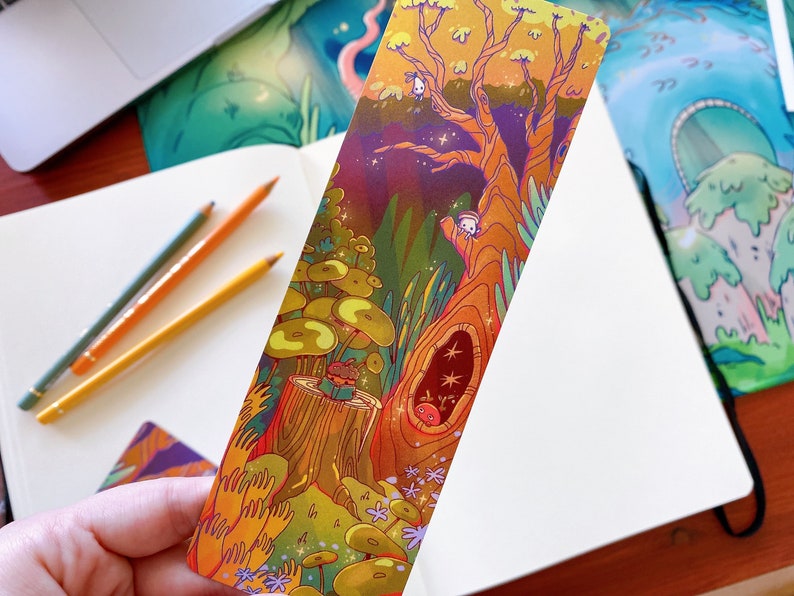 May include: A colorful bookmark featuring a whimsical illustration of a tree with a hollow trunk, a small creature peeking out, and a mushroom forest. The bookmark is printed on thick cardstock and has a glossy finish.