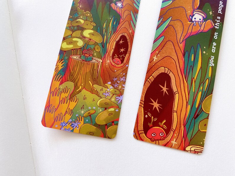 May include: Two colorful bookmarks with whimsical illustrations of a forest scene. The bookmarks feature a tree trunk with hollow spaces, mushrooms, and small creatures. One bookmark has the text "you are on this page" on the right side.