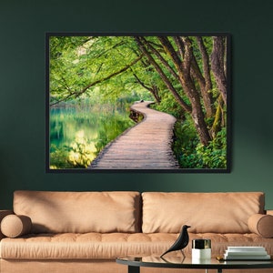 May include: A rectangular wall art print featuring a wooden boardwalk winding through a lush green forest beside a calm lake. The print is displayed in a black frame above a tan leather sofa, a glass coffee table, and a tall floor lamp.
