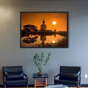 May include: A framed wall art print depicting a large Buddha statue silhouette against a vibrant orange sunset, reflected in a calm body of water. The scene includes a dark tree silhouette and clear sky, presented in a black frame on a gray wall.