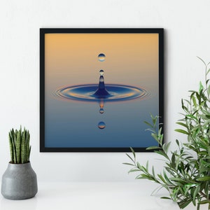 May include: A square wall art print in a thin black frame featuring a high-speed photograph of a water droplet impact. The image displays a central blue water crown with concentric ripples, reflecting a gradient of soft orange and deep blue.