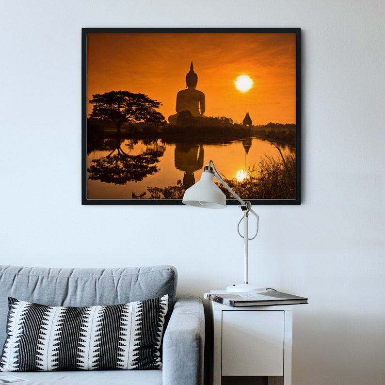 May include: A framed wall art print depicting a silhouette of a large Buddha statue at sunset, reflected in a calm body of water. The scene features warm orange tones, a dark tree, and a bright sun, displayed above a gray sofa and a white lamp.