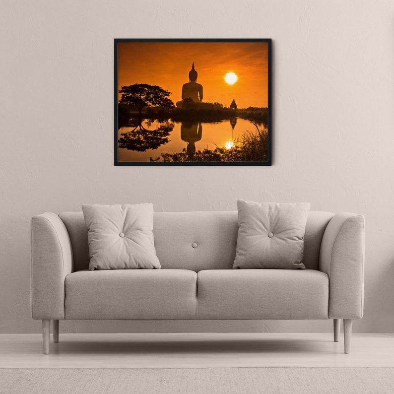 May include: A framed wall art print featuring a silhouette of a large Buddha statue at sunset, with the orange sun and statue reflected in a calm body of water. The scene includes a dark tree on the left and a clear, vibrant orange sky background.