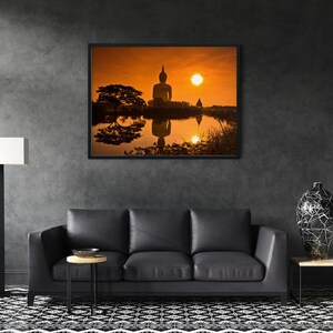 May include: A rectangular wall art print featuring a silhouette of a large Buddha statue at sunset, with the sun and statue reflected in a calm body of water. The scene includes a dark tree on the left and a clear orange sky, framed in a thin black border.