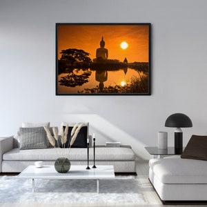 May include: A framed wall art print featuring a silhouette of a seated Buddha statue against a vibrant orange sunset. The scene includes a large tree and a small structure reflected in the calm water below, all captured in a horizontal landscape.