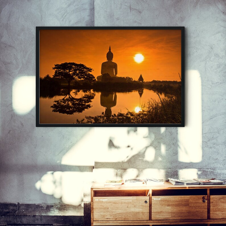 May include: A framed wall art print featuring a silhouette of a seated Buddha statue at sunset. The orange sky and sun are reflected in the calm water below, alongside a large tree. The artwork is displayed in a thin black frame on a gray wall.