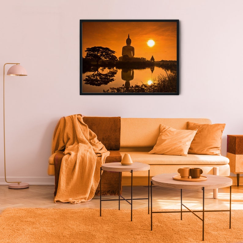 May include: A framed wall art print depicting a silhouette of a Buddha statue at sunset over a reflective body of water, displayed above a light orange sofa with a matching throw blanket, two round side tables, and a tall floor lamp.